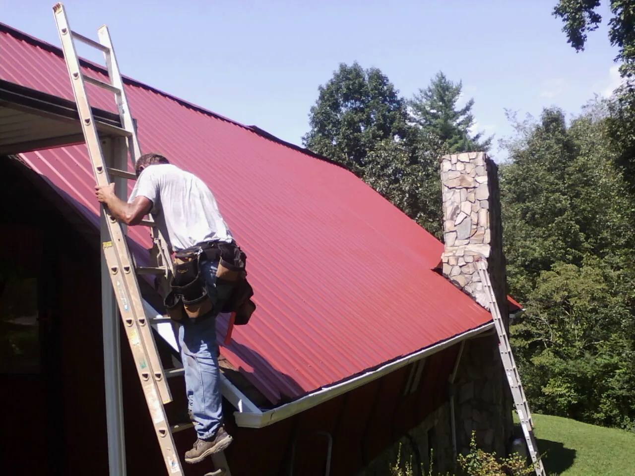 Roofer on a ladder beside a red metal roof and stone chimney — Soffit & Fascia Repair by Roofing Brothers in Crater Lake